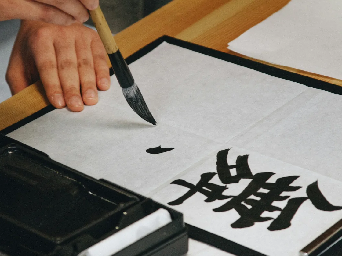 Mano trazando un signo con un pincel cargado de tinta negra sobre papel claro, junto a un tintero plano